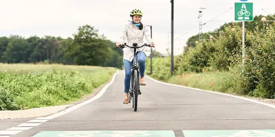 Person mit weißer Jacke und Helm fährt auf einem Fahrradweg, umgeben von Grünflächen und Verkehrsschildern für Radfahrer.