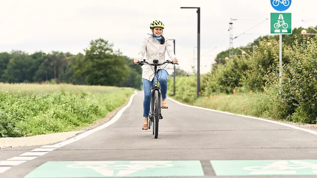 Person mit weißer Jacke und Helm fährt auf einem Fahrradweg, umgeben von Grünflächen und Verkehrsschildern für Radfahrer.