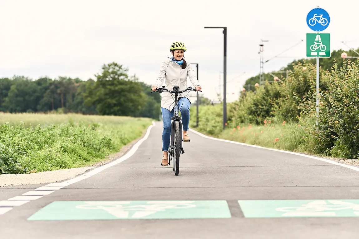 Person mit weißer Jacke und Helm fährt auf einem Fahrradweg, umgeben von Grünflächen und Verkehrsschildern für Radfahrer.