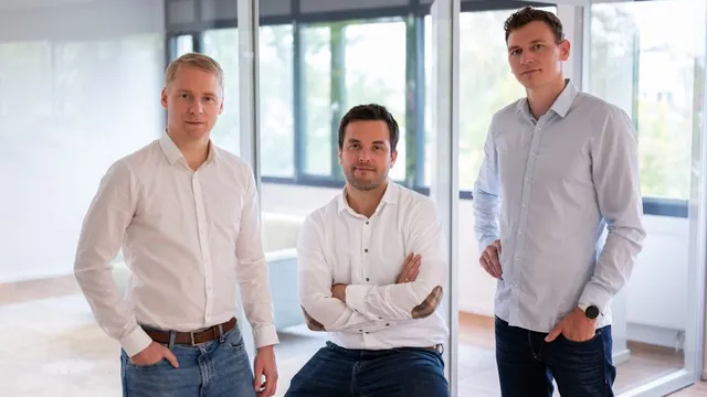 Drei Männer in Hemden und Jeans in einem modernen Büro mit Glaswänden, einer sitzt auf einem Hocker, zwei stehen.