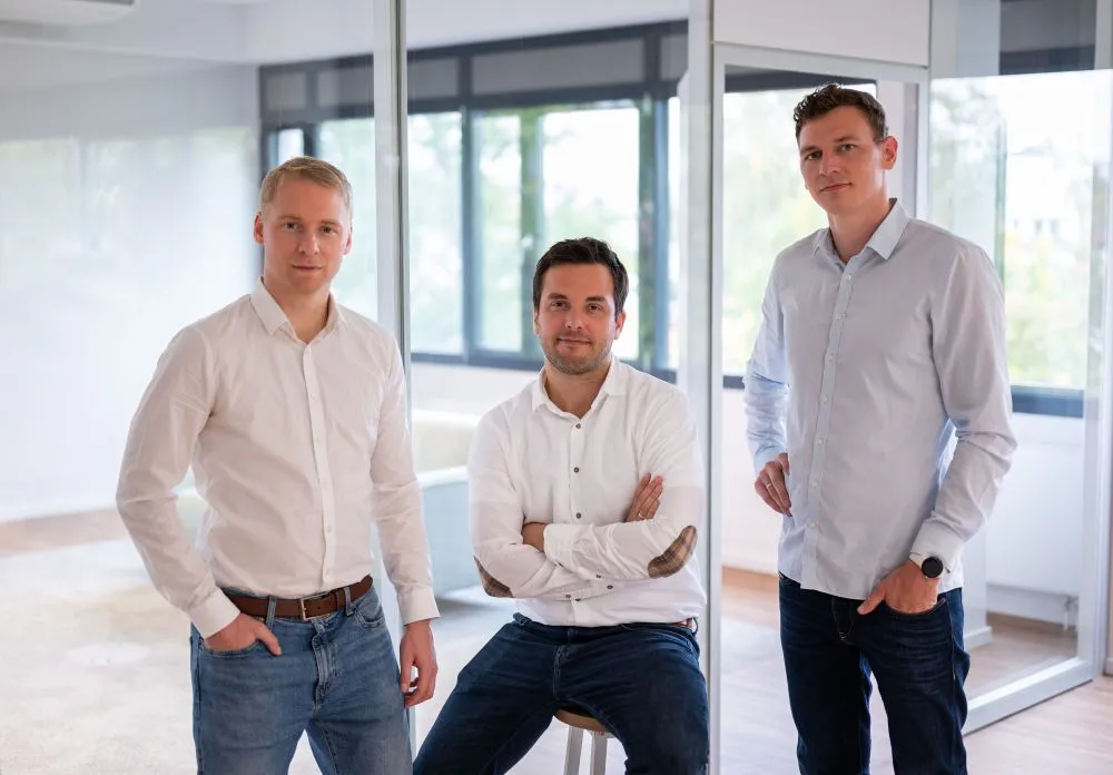 Drei Männer in Hemden und Jeans in einem modernen Büro mit Glaswänden, einer sitzt auf einem Hocker, zwei stehen.