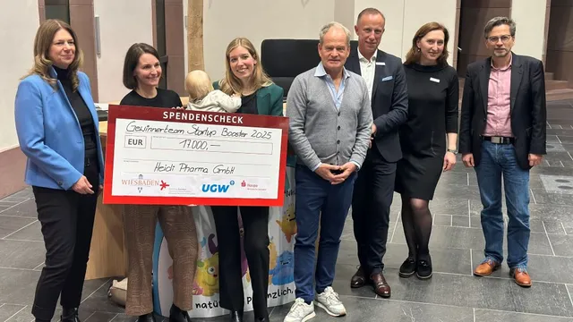 Gruppe von sieben Personen steht in einem Innenraum, zwei halten einen großen Spendenscheck mit der Aufschrift 'Gutscheinteam Startup Booster 2025' und 'Heidi Pharma GmbH'.