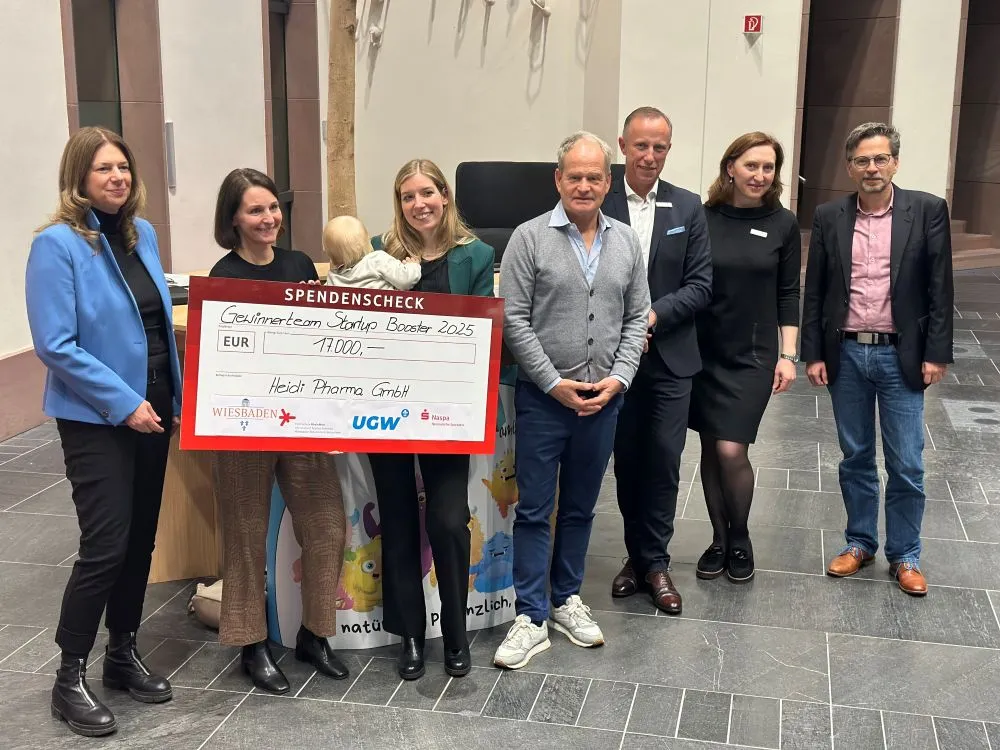 Gruppe von sieben Personen steht in einem Innenraum, zwei halten einen großen Spendenscheck mit der Aufschrift 'Gutscheinteam Startup Booster 2025' und 'Heidi Pharma GmbH'.