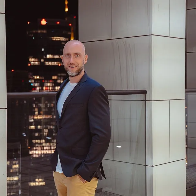 Ein Mann mit Sakko und hellen Hosen steht vor einer Glasbrüstung auf einer Terrasse mit Blick auf beleuchtete Hochhäuser bei Nacht