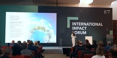 Konferenzsaal mit Publikum und einer Person auf der Bühne vor einer Leinwand und einem großen Schriftzug 'INTERNATIONAL IMPACT FORUM'