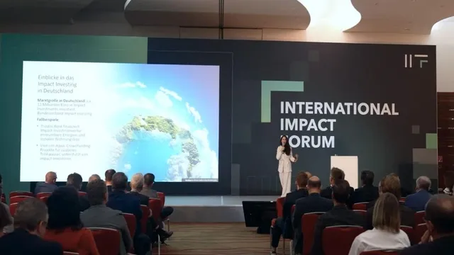 Konferenzsaal mit Publikum und einer Person auf der Bühne vor einer Leinwand und einem großen Schriftzug 'INTERNATIONAL IMPACT FORUM'