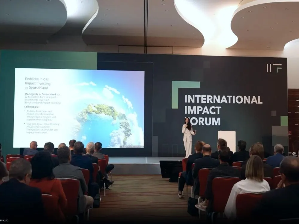 Konferenzsaal mit Publikum und einer Person auf der Bühne vor einer Leinwand und einem großen Schriftzug 'INTERNATIONAL IMPACT FORUM'