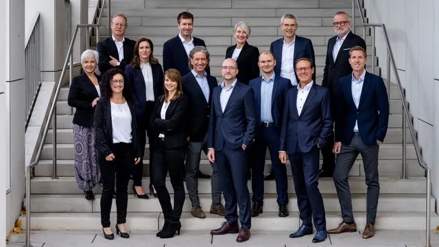 Gruppe von 15 Personen in Businesskleidung steht auf einer breiten Treppe in mehreren Reihen