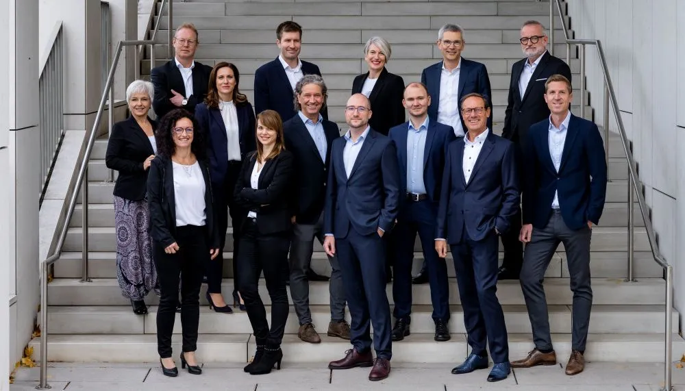 Gruppe von 15 Personen in Businesskleidung steht auf einer breiten Treppe in mehreren Reihen