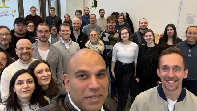 Gruppe von Menschen in einem Innenraum, einige tragen Pullover und Hemden, im Hintergrund sind Fenster und eine Wand mit Bildern zu sehen.
