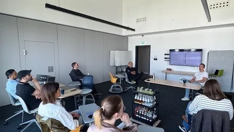 Mehrere Personen sitzen in einem modernen Besprechungsraum auf Stühlen mit gelben und grauen Sitzflächen und hören einem Vortragenden zu, der vor einem Bildschirm steht.