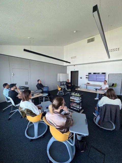 Mehrere Personen sitzen in einem modernen Besprechungsraum auf Stühlen mit gelben und grauen Sitzflächen und hören einem Vortragenden zu, der vor einem Bildschirm steht.
