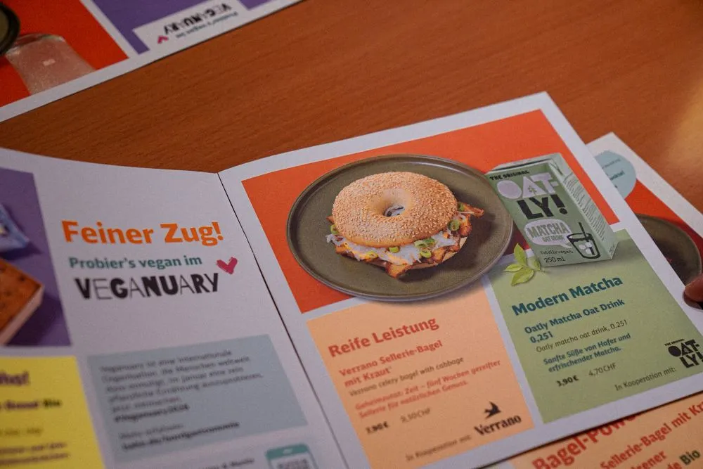 Aufgeschlagene Broschüre mit einem Bagel auf einem Teller und Texten zu veganem Angebot und Matcha-Getränk
