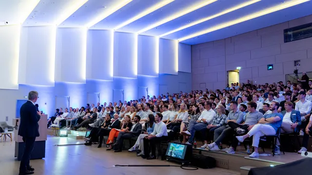 Vortragender steht vor großem Publikum in modernem Auditorium mit beleuchteter Decke