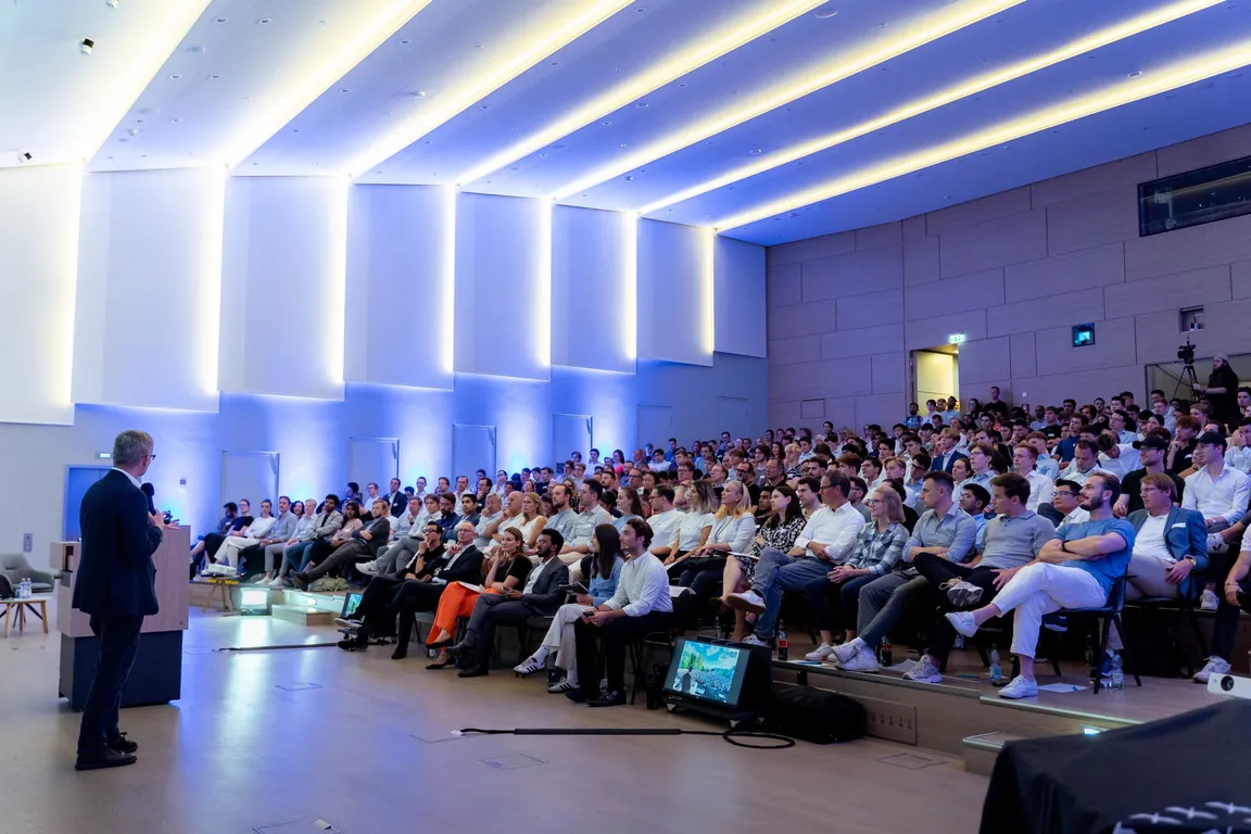 Vortragender steht vor großem Publikum in modernem Auditorium mit beleuchteter Decke