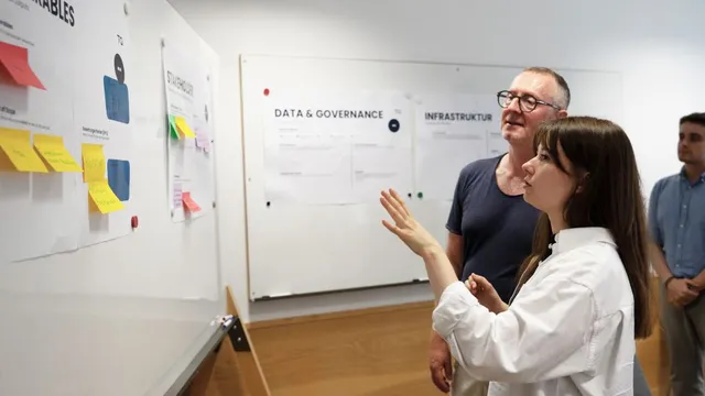 Zwei Personen stehen vor einer Wand mit mehreren angebrachten Zetteln und Diagrammen, eine Person zeigt auf die Wand, im Hintergrund weitere Diagramme mit den Überschriften 'DATA & GOVERNANCE' und 'INFRASTRUKTUR'
