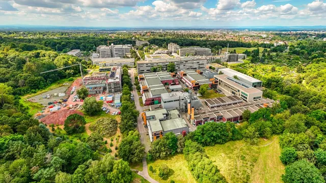 Luftaufnahme eines Campus mit mehreren Gebäuden, umgeben von viel Grün und Bäumen, im Hintergrund eine Stadtlandschaft
