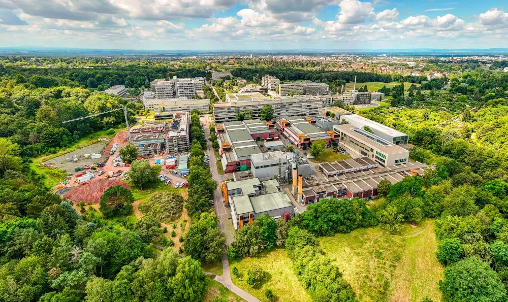 Luftaufnahme eines Campus mit mehreren Gebäuden, umgeben von viel Grün und Bäumen, im Hintergrund eine Stadtlandschaft