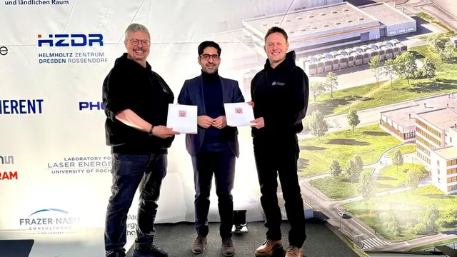 Drei Personen stehen vor einer Wand mit Logos und einem großen Gebäudeplan, zwei halten jeweils ein Dokument in der Hand.