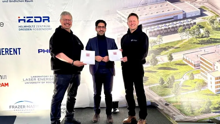 Drei Personen stehen vor einer Wand mit Logos und einem großen Gebäudeplan, zwei halten jeweils ein Dokument in der Hand.