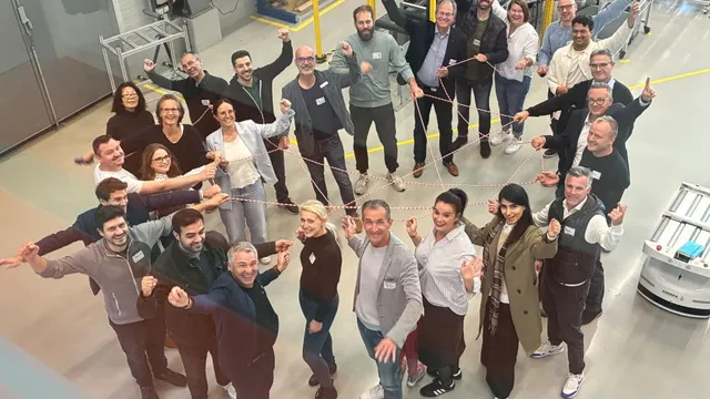 Gruppe von Menschen steht in einer Halle im Kreis und zeigt mit ausgestrecktem Arm nach innen