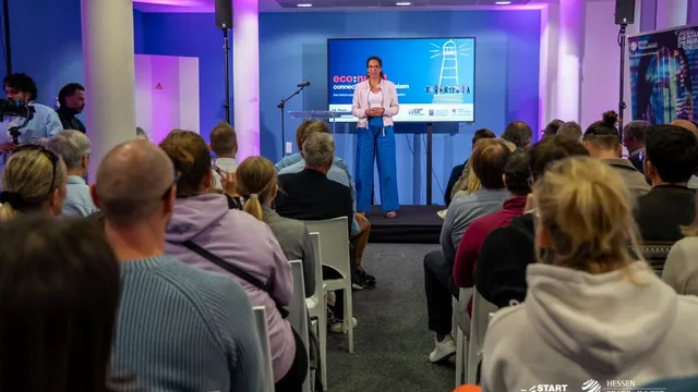 Frau steht auf Bühne vor Publikum, im Hintergrund Bildschirm mit Text 'econnect' und Logos von Start Hub Hessen und Hessen Trade & Invest.