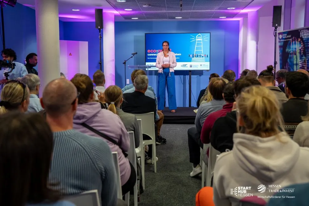 Frau steht auf Bühne vor Publikum, im Hintergrund Bildschirm mit Text 'econnect' und Logos von Start Hub Hessen und Hessen Trade & Invest.