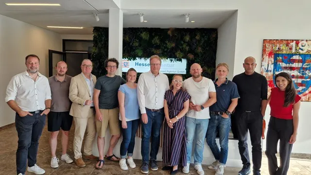 Gruppe von elf Personen steht in einem Raum vor einer Wand mit Pflanzen und einem Bildschirm mit dem Text 'Hessen Agentur GmbH' beim Kickoff-Event Slush 2025