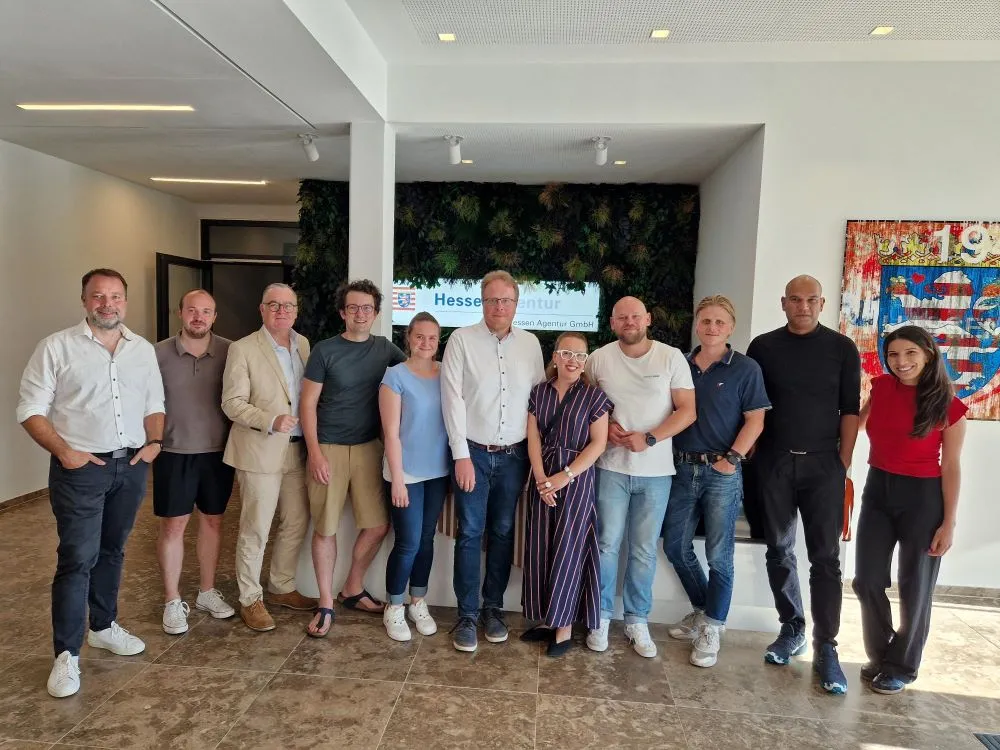 Gruppe von elf Personen steht in einem Raum vor einer Wand mit Pflanzen und einem Bildschirm mit dem Text 'Hessen Agentur GmbH' beim Kickoff-Event Slush 2025