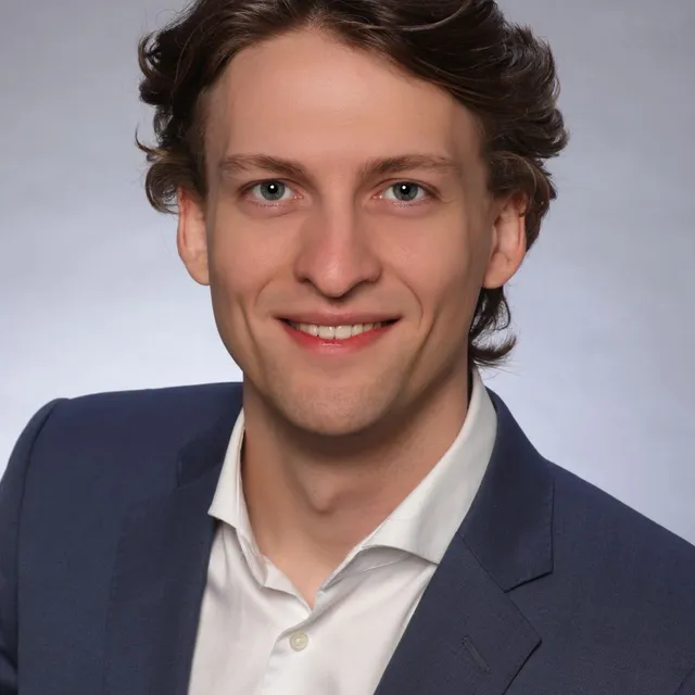 Mann mit gewelltem Haar trägt weißes Hemd und dunklen Anzug vor neutralem Hintergrund