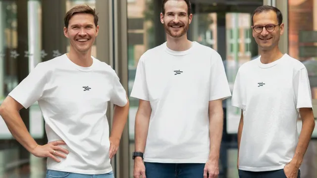 Drei Männer in weißen T-Shirts mit kleinem schwarzem Logo stehen nebeneinander in einem hellen Innenraum und lächeln in die Kamera