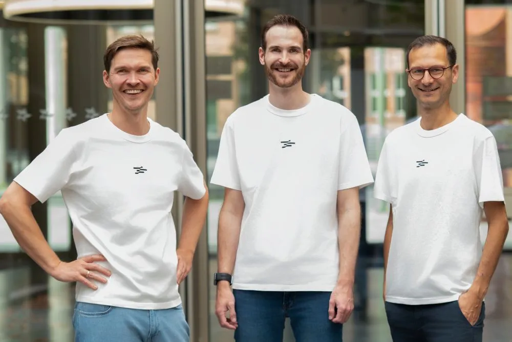 Drei Männer in weißen T-Shirts mit kleinem schwarzem Logo stehen nebeneinander in einem hellen Innenraum und lächeln in die Kamera
