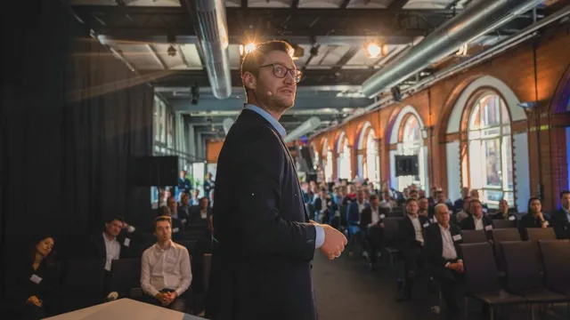 Mann im Anzug steht vor Publikum in einem großen Raum mit hohen Fenstern und sichtbaren Deckenrohren