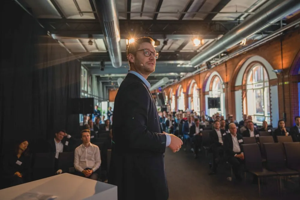 Mann im Anzug steht vor Publikum in einem großen Raum mit hohen Fenstern und sichtbaren Deckenrohren