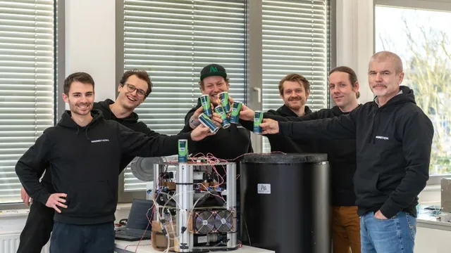 Fünf Personen in schwarzer und blauer Kleidung stehen um technische Geräte und stoßen Dosen an.