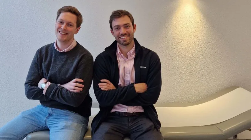 Zwei Männer sitzen mit verschränkten Armen auf einer grauen Liege vor einer weißen Wand.