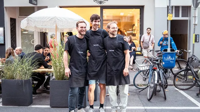 Drei Personen mit schwarzen Schürzen stehen vor einem Laden mit dem Schriftzug 'BioKaiser' und posieren vor einem Tisch mit Gästen und mehreren Fahrrädern