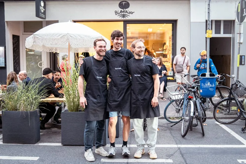 Drei Personen mit schwarzen Schürzen stehen vor einem Laden mit dem Schriftzug 'BioKaiser' und posieren vor einem Tisch mit Gästen und mehreren Fahrrädern