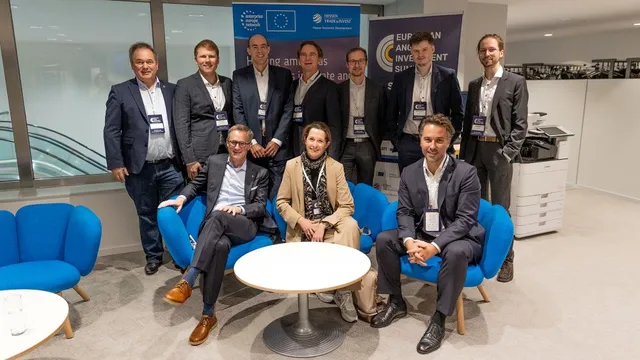Neun Personen in Businesskleidung posieren in einem modernen Büro mit blauen Sesseln und Bannern im Hintergrund.