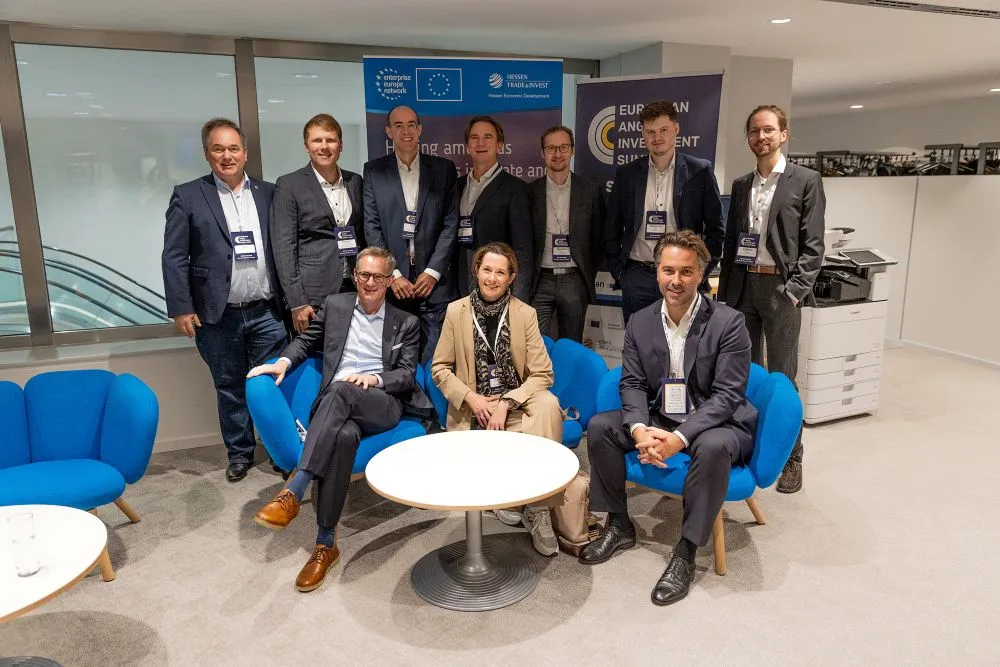 Neun Personen in Businesskleidung posieren in einem modernen Büro mit blauen Sesseln und Bannern im Hintergrund.