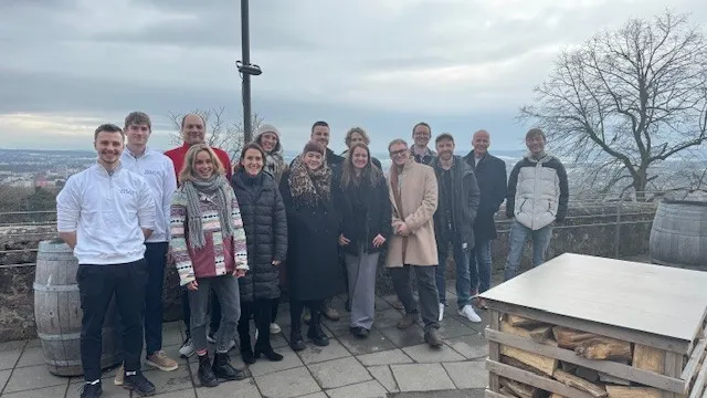 Gruppe von Menschen steht auf gepflastertem Platz mit Weinfässern und Holzstapel im Hintergrund unter bewölktem Himmel