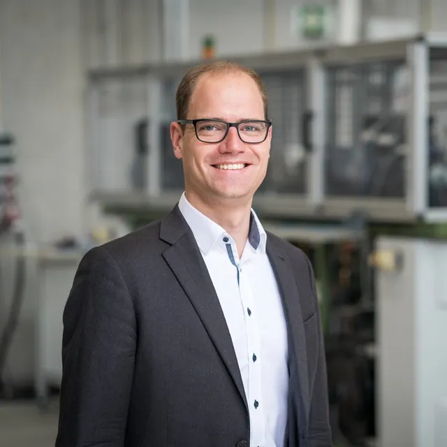 Mann mit Brille, weißem Hemd und dunklem Sakko lächelt vor unscharfem industriellem Hintergrund