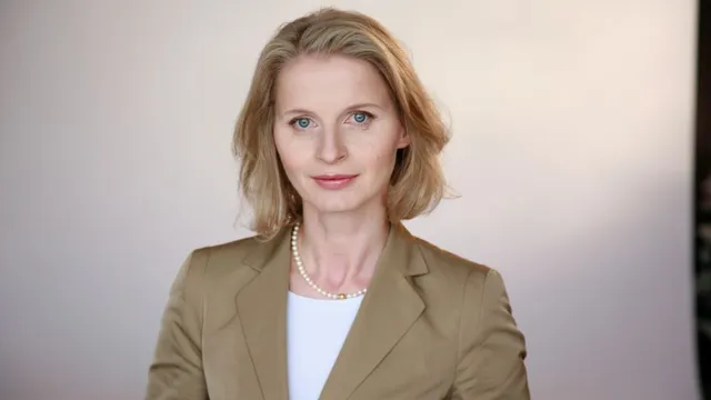 Frau mit schulterlangem blonden Haar trägt eine beige Jacke, weiße Bluse und Perlenkette vor neutralem Hintergrund