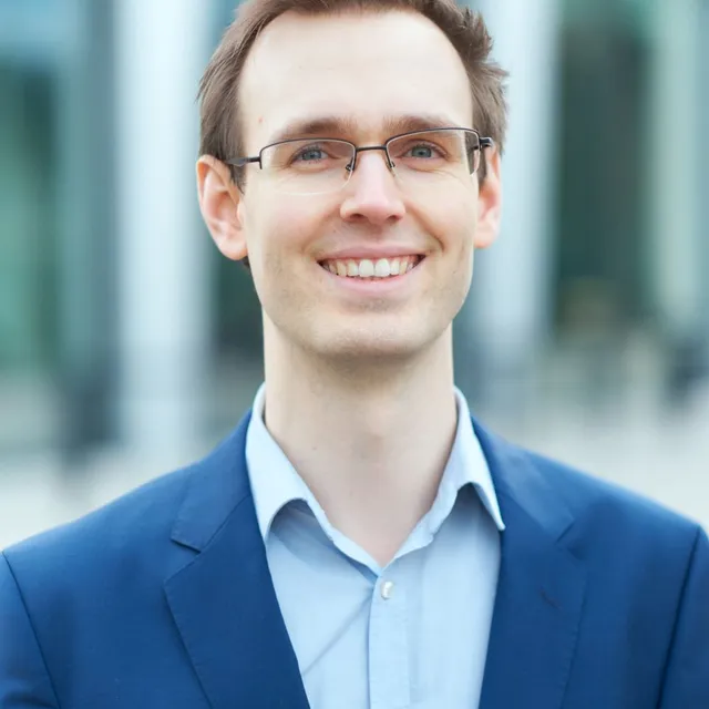 Junger Mann in blauem Sakko und hellblauem Hemd vor unscharfem Gebäudehintergrund