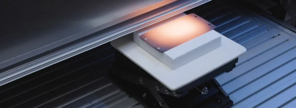 Nahaufnahme eines weißen, quadratischen Geräts mit beleuchteter Oberfläche auf einer schwarzen, höhenverstellbaren Plattform vor einem Aluminiumprofil-Hintergrund