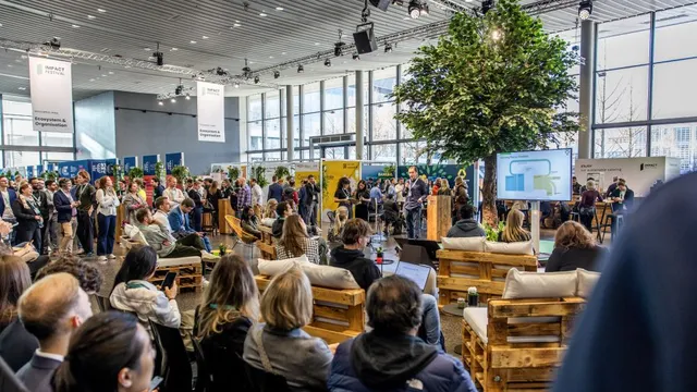 Große Gruppe sitzender Menschen bei einer Veranstaltung in hellem Raum mit großen Fenstern, Holzbankreihen und einem Baum in der Mitte.