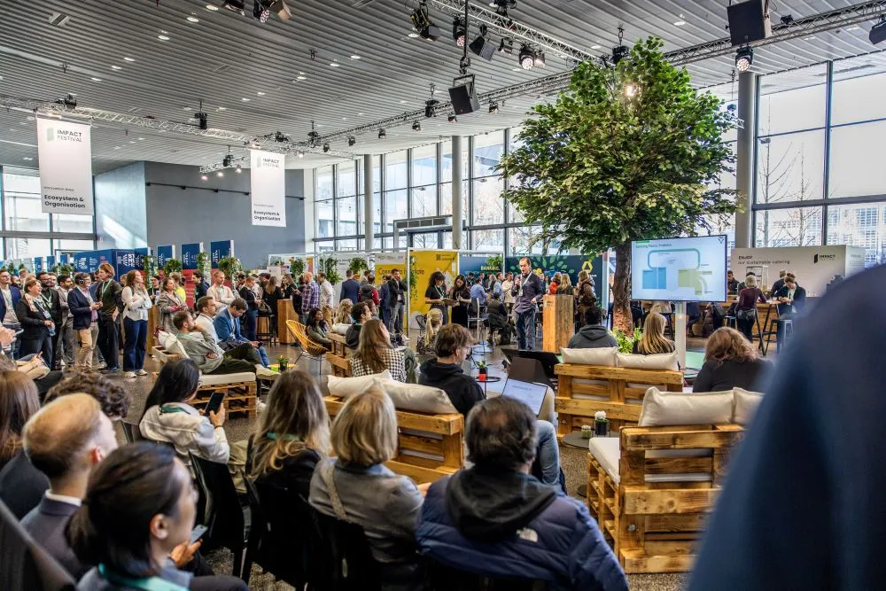 Große Gruppe sitzender Menschen bei einer Veranstaltung in hellem Raum mit großen Fenstern, Holzbankreihen und einem Baum in der Mitte.