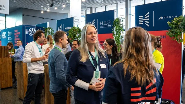 Mehrere Personen stehen in einem hellen Messebereich mit blauen und roten Stellwänden, auf denen "Start Hub" und "CESAH" steht. Einige tragen Namensschilder und grüne Lanyards.
