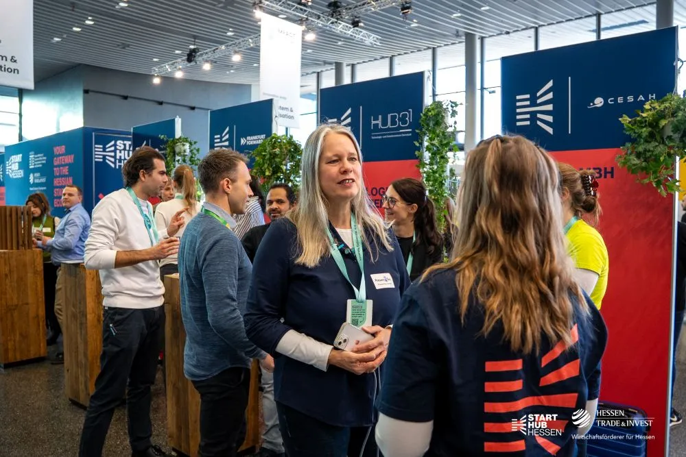 Mehrere Personen stehen in einem hellen Messebereich mit blauen und roten Stellwänden, auf denen "Start Hub" und "CESAH" steht. Einige tragen Namensschilder und grüne Lanyards.