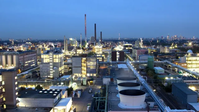 Industriepark mit beleuchteten Fabrikgebäuden, hohen Schornsteinen und großen Lüftungstürmen bei Dämmerung.